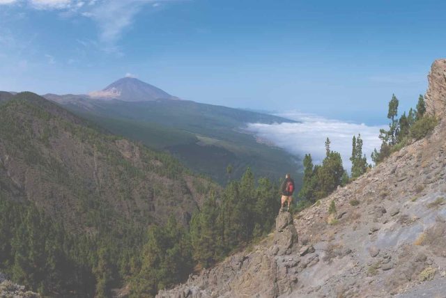 A hiker stands on a rock peak and looks out over the vast mountain landscape of Tenerife. In the distance is the highest mountain in Spain. Below right are clouds.