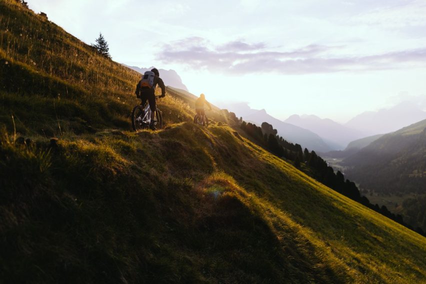 Due rider MTB su sentiero erboso al tramonto, tra colline stratificate e luce dorata